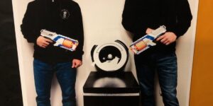 A Photo Of Two Men From The Shoulders Down Standing And Pointing Nerf Guns At A Roomba On A Pedestal Between Them.jpg