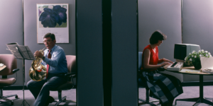 A Photo Showing Two People In Separate Cubicles One Is Playing A French Horn And The Other Is Typing At A Computer.png