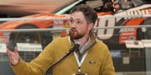 Man In A Yellow Sweater Standing At A Podium Speaking Into A Microphone Info For Editor If Needed.jpg