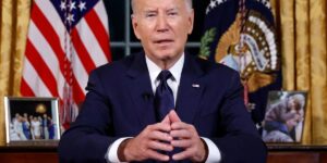 President Joe Biden Sitting At A Desk With Hands Together Talking To The Camera.jpg