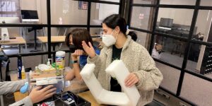 2 People With Masks On Sitting In At A Desk Chatting With Various Different Boxes And Wires Around Them.jpg