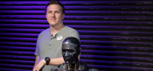 A White Man Standing Alongside A Human Looking Robot Seated In A Red Chair.png