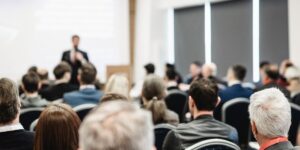 Rear View Of People Sitting In Chairs Looking At Someone Speaking In Background.jpg