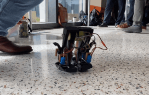 An Animated Gif Showing A Four Inch Tall Robot That Consists Only Of Two Legs Walking Jauntily Across A Conference Floor.gif