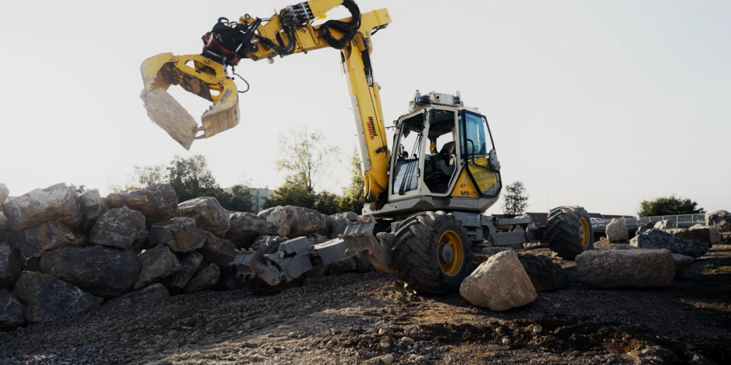 An Excavator With No Human Inside And Sensors On Its Roof Picks Up A Large Rock And Places It On A Partially Completed Rock Wall.png