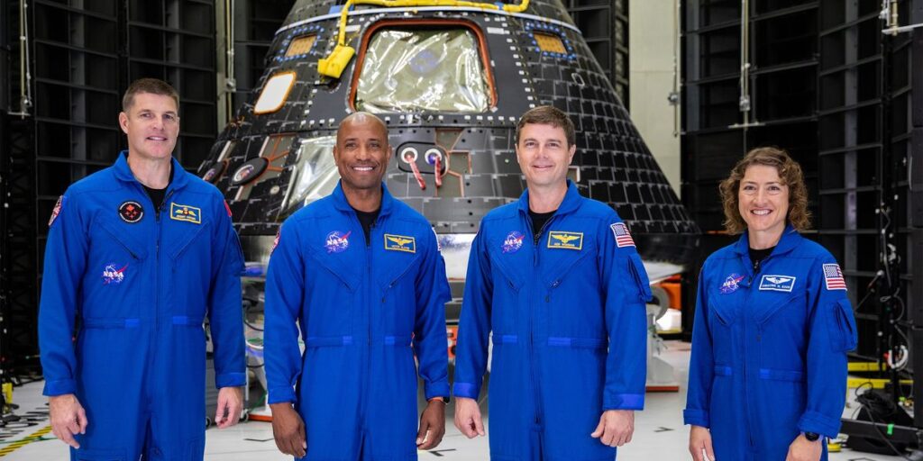 Three Men And A Woman All Dressed In Blue Uniforms Stand Abreast And In Front Of A Large Semiconical Space Capsule.jpg