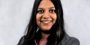 Woman With Long Dark Hair Wearing A Pink Button Up Shirt And A Gray Blazer Smiling At The Camera Against A Light Background.jpg