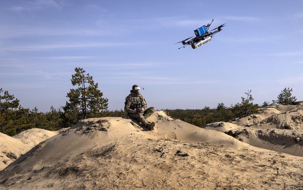 a soldier sits on a sandy hill wearing special glasses and holding a remote to control a drone with